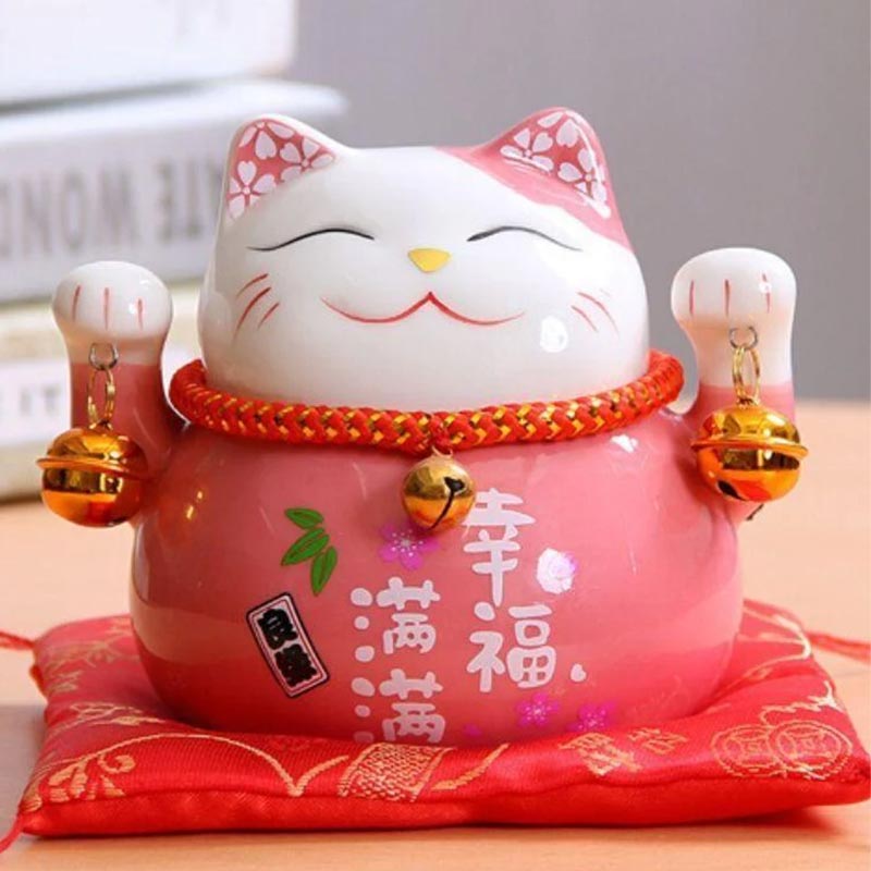 Un chat maneki neko rose posé sur un coussin rouge. La statuette chat japonais tient des clochette. Cet objet deco japonais est en céramique et peint à la main avec de la peinture acrylique