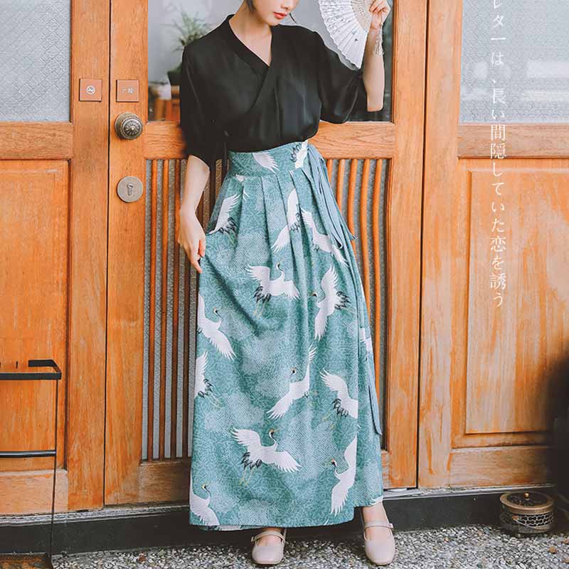 Une femme est habillé avec un ensemble pantalon hakama. Le haut en forme de kimono est de couleur noir et la robe kimono est imprimé avec le motif japonais de la grue Tsuru