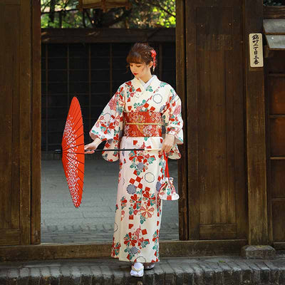 Une femme est habillée avec un kimono japonais traditionnel femme soie. La Robe kimono de tradition japonaise, ornée de motifs floraux sakura et fukiwa sur un tissu blanc. La femme se tient debout avec une ombrelle japonaise. Son kimono est fermé avec une ceinture obi rouge