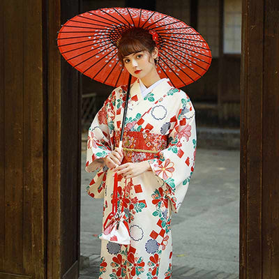 Une femme porte un kimono traditionnel japonais femme de couleur blanche. Son kimonot japonais est fermé avec une ceinture obi roge. Elle tient dans la main une ombrelle japonaise de couleur rouge et fabriqué en papier